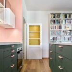 Kuchynská ulička medzi tmavozelenými skrinkami s ružovými úchytmi, biele dvere so žltým sklom a biela knižnica v pozadí.
