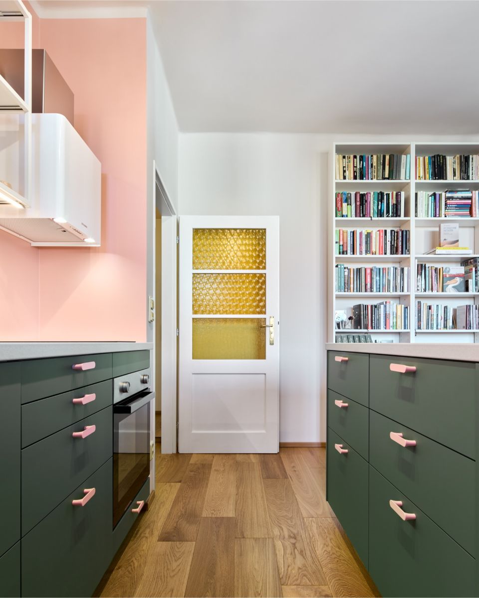 Kuchynská ulička medzi tmavozelenými skrinkami s ružovými úchytmi, biele dvere so žltým sklom a biela knižnica v pozadí.