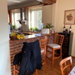 Starší kuchynský bar s tehlovým obkladom, drevené barové stoličky a biela dlažba v pôvodnom interiéri.