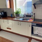 Staršia biela kuchyňa s nerezovým drezom, zabudovanou rúrou a malým štvorcovým obkladom na stene.