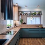 Rohová modrozelená kuchyňa s dubovým dekorom, čiernym drezom a policou s prevísajúcimi zelenými rastlinami.