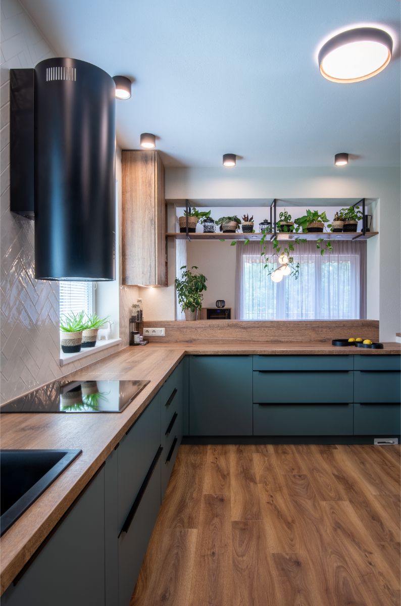 Rohová modrozelená kuchyňa s dubovým dekorom, čiernym drezom a policou s prevísajúcimi zelenými rastlinami.