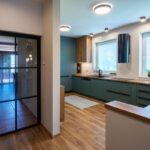 Presklené dvere s čiernym rámom vedľa modernej kuchynskej linky v petrolejovej farbe s drevenými detailmi.