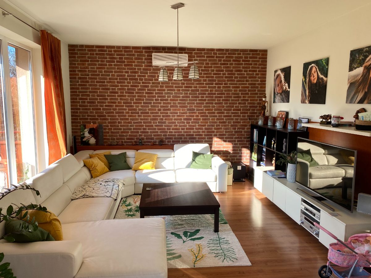 Pôvodná obývacia izba s tehlovou stenou, bielou rohovou sedačkou a tmavým konferenčným stolíkom pred renováciou.