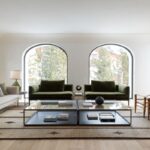 Presvetlená obývacia izba s dvoma zelenými zamatovými kreslami, veľkými oblúkovými oknami a dvojitým skleneným stolíkom na koberci.