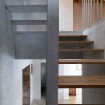 Moderné interiérové schodisko s drevenými dubovými stupňami, sivou betónovou stenou a kovovým zábradlím nad kamienkovou dlažbou.