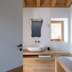 Kúpeľňa so svetlým dreveným obkladom, bielym umývadlom na doske, asymetrickým zrkadlom a priznanými stropnými trámami.