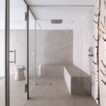 Minimalistická sauna s béžovým obkladom, sklenenými dverami, kamennou lavicou a dekoratívnymi kmeňmi briez.