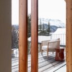 Drevený balkón s kovovým zábradlím, svetlým pleteným kreslom a hnedým keramickým stolíkom s výhľadom na zasnežené hory.
