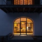 Osvetlený interiér jedálne s čiernym stolom a drevenými stoličkami videný cez veľké oblúkové okno v tmavej fasáde.