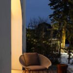 Podsvietený výklenok bielej steny s dizajnovým pleteným kreslom a hnedým lesklým stolíkom na tmavej drevenej terase.