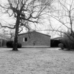 Pôvodný tehlový dom so sedlovou strechou a malým bielym oknom v zatrávnenej záhrade s listnatým stromom.