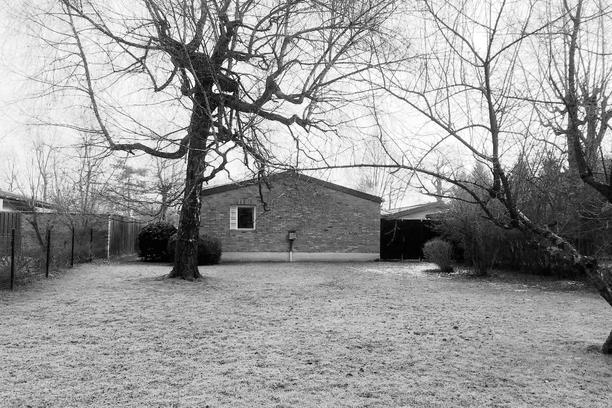 Pôvodný tehlový dom so sedlovou strechou a malým bielym oknom v zatrávnenej záhrade s listnatým stromom.