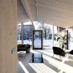 Presvetlená obývacia izba s dreveným obkladom, skleneným štítom a dizajnovými kreslami na svetlej podlahe.