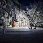 Osvetlený dom v tme s preskleným priečelím a tehlovými stenami uprostred zasneženej záhrady.