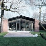 Zmodernizovaný dom s presklenou fasádou s čiernym rámom, červeným tehlovým obkladom a svetlou kamennou terasou.