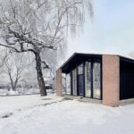 Zrekonštruovaný dom s tehlovými múrmi a veľkým presklením v čiernych rámoch v zasneženej krajine.