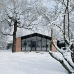 Zrekonštruovaný dom s tehlovými múrmi a veľkým presklením v čiernych rámoch v zasneženej krajine.