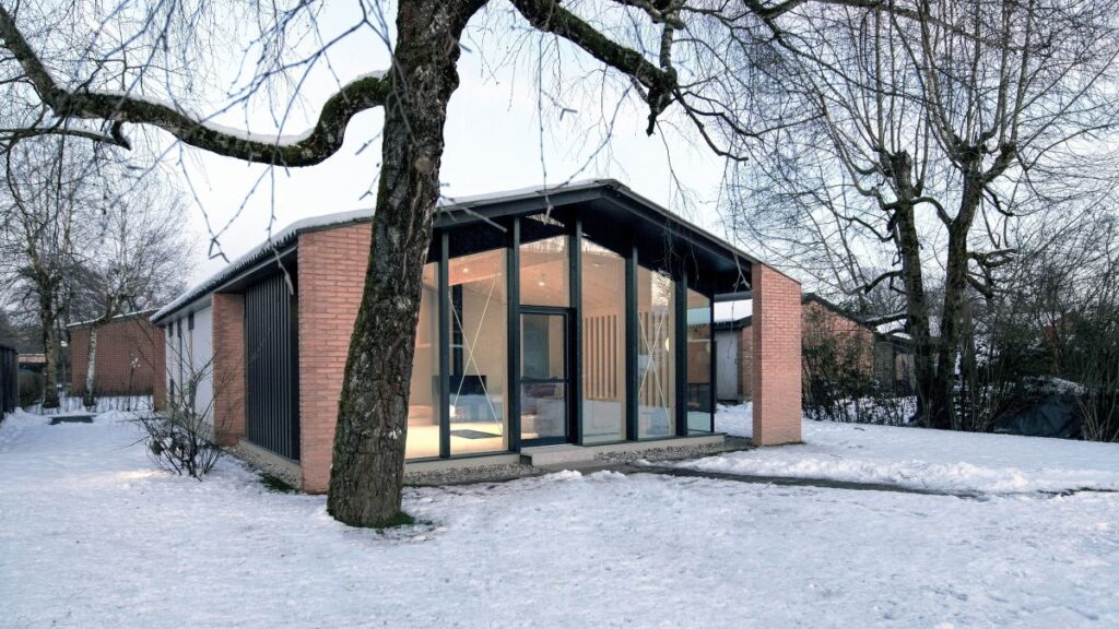 Dom s tehlovými múrmi a veľkým presklením v čiernych rámoch v zasneženej krajine.