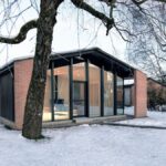 Dom s tehlovými múrmi a veľkým presklením v čiernych rámoch v zasneženej krajine.