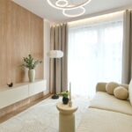 Presvetlená izba s dreveným stenovým panelom, nízkou béžovou komodou, svetlou pohovkou a oblúkovým stropným svietidlom v japandi štýle.