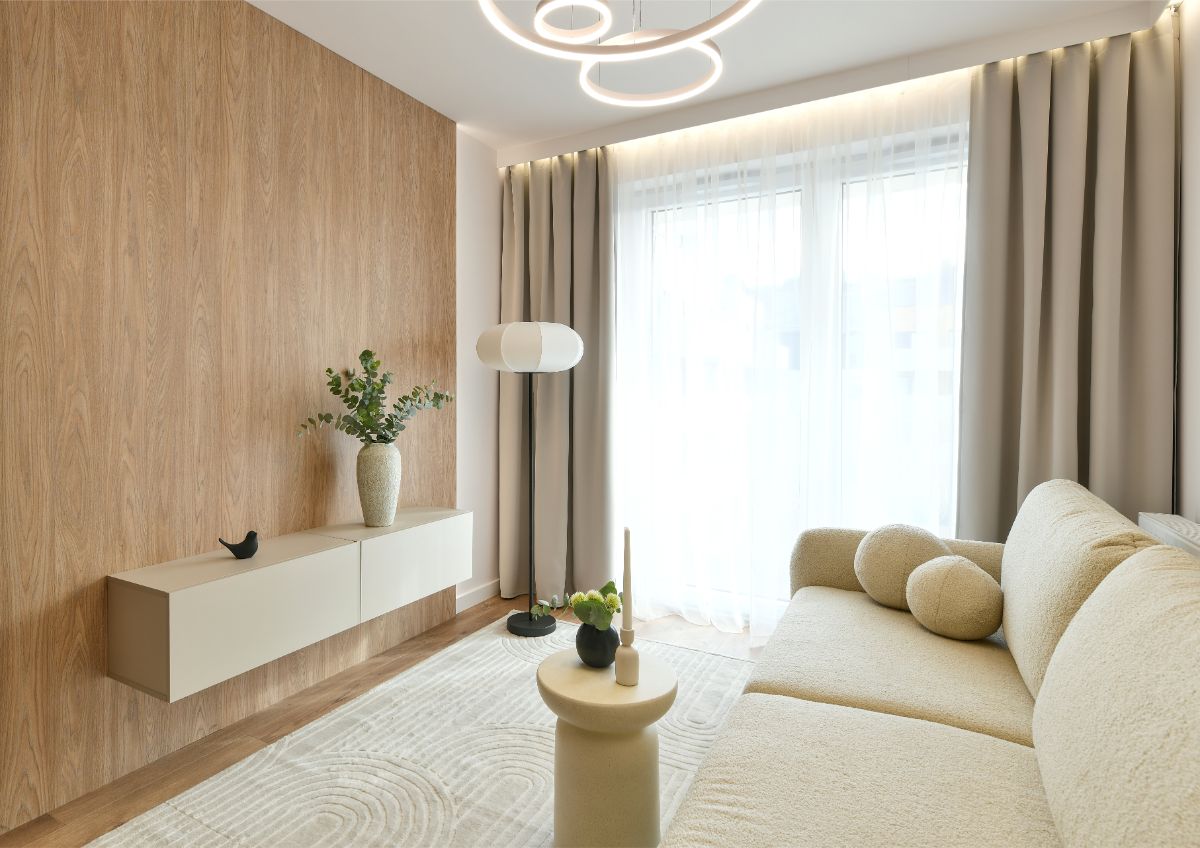 Presvetlená izba s dreveným stenovým panelom, nízkou béžovou komodou, svetlou pohovkou a oblúkovým stropným svietidlom v japandi štýle.