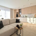 Otvorený priestor s bledou kuchynskou linkou, dreveným dekorom, sivou sedačkou a prírodným tkaným kobercom v japandi koncepte.