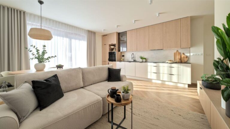 Otvorený priestor s bledou kuchynskou linkou, dreveným dekorom, sivou sedačkou a prírodným tkaným kobercom v japandi koncepte.