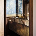 Toaleta s kamenným umývadlom na skrinke v mosadznej farbe a tmavomodrou tapetou s rastlinným vzorom.