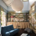 Detská izba s modrou čalúnenou sedačkou, maľovanými stenami motívom prírody a otvorenou policou.