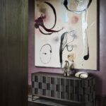 Chodba s abstraktným obrazom na fialovej stene nad tmavou drevenou komodou..