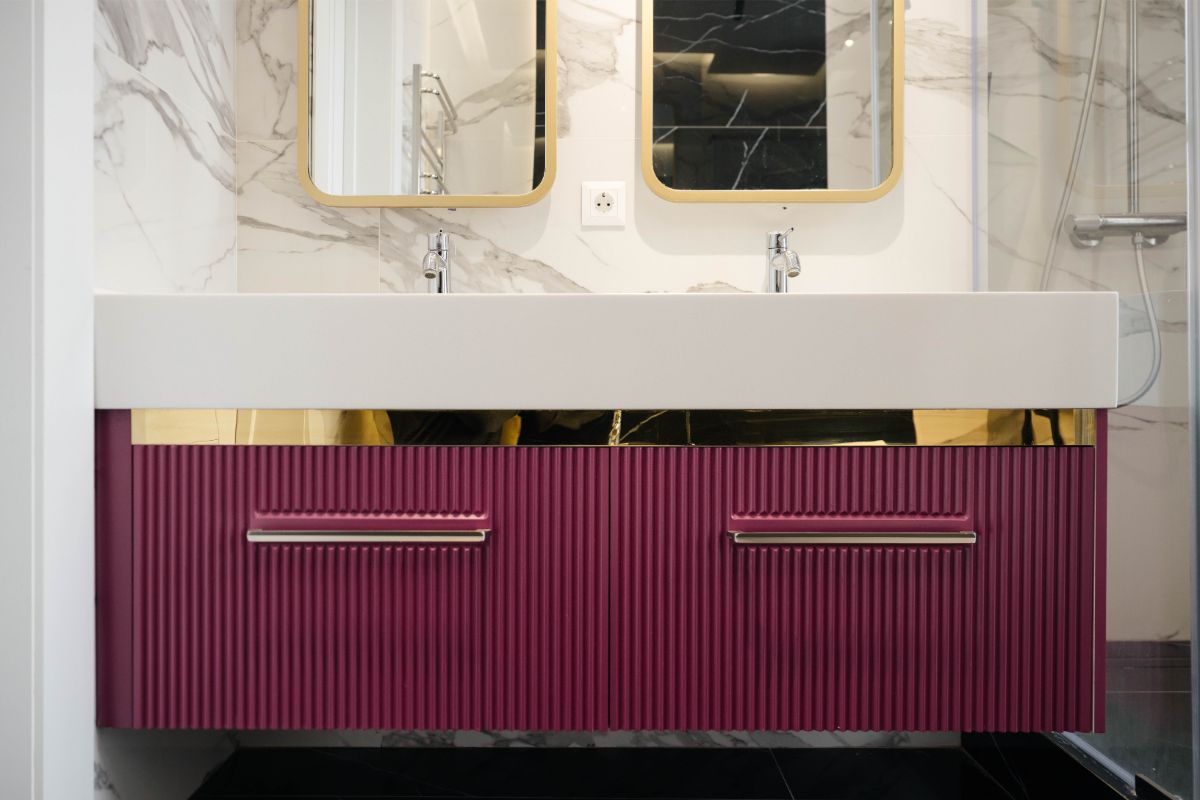 Bordová drážkovaná skrinka so zlatou lištou, biele mramorové obklady a hranaté zrkadlá v tenkom zlatom ráme.