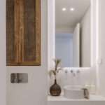 Minimalistické biele umývadlo na doske, nástenná chrómová batéria, podsvietené zrkadlo a drevená skrinka s ratanovým výpletom.