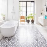 Presvetlený interiér s výraznou vzorovanou dlažbou, bielou vaňou, čiernymi kovovými dverami a živými zelenými rastlinami.