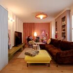 Moderná obývacia izba s hnedou zamatovou sedačkou, žltou taburetkou, ružovými závesmi a drevenou podlahou.