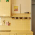 Žltá kachličková kuchynská linka s antikorovou varnou doskou, sklenenými koreničkami a zeleným lineárnym svietidlom.