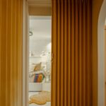 Skladajúce posuvné dvere v žltej farbe, zakrývajúce spálňu s posteľou a nabielo natretou tehlovou stenou.