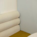 Čalúnená lavica s olivovou textíliou, bielym valcovitým operadlom a svetlomodrou stenou v minimalistickom detaile.