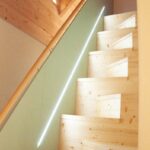 Drevené lomené schodisko so zabudovaným líniovým LED podsvietením v svetlozelenej bočnej stene pod šikmým stropom.