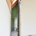 Úzka biela chodba s betónovou dlažbou, zelenými dverami a čiernym kovovým rebríkom vedúcim k strešnému oknu.