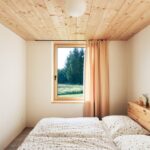 Minimalistická spálňa s nízkym dreveným stropom, manželskou posteľou pod oknom a guľatým bielym stropným svietidlom.