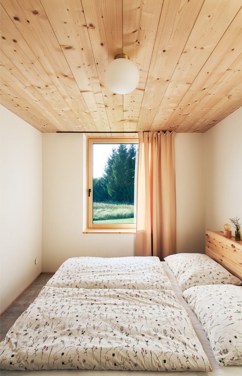 Minimalistická spálňa s nízkym dreveným stropom, manželskou posteľou pod oknom a guľatým bielym stropným svietidlom.