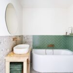 Moderná kúpeľňa s bielou vaňou, zeleným obkladom v tvare rybej kosti a dreveným stolíkom pod oválnym umývadlom.
