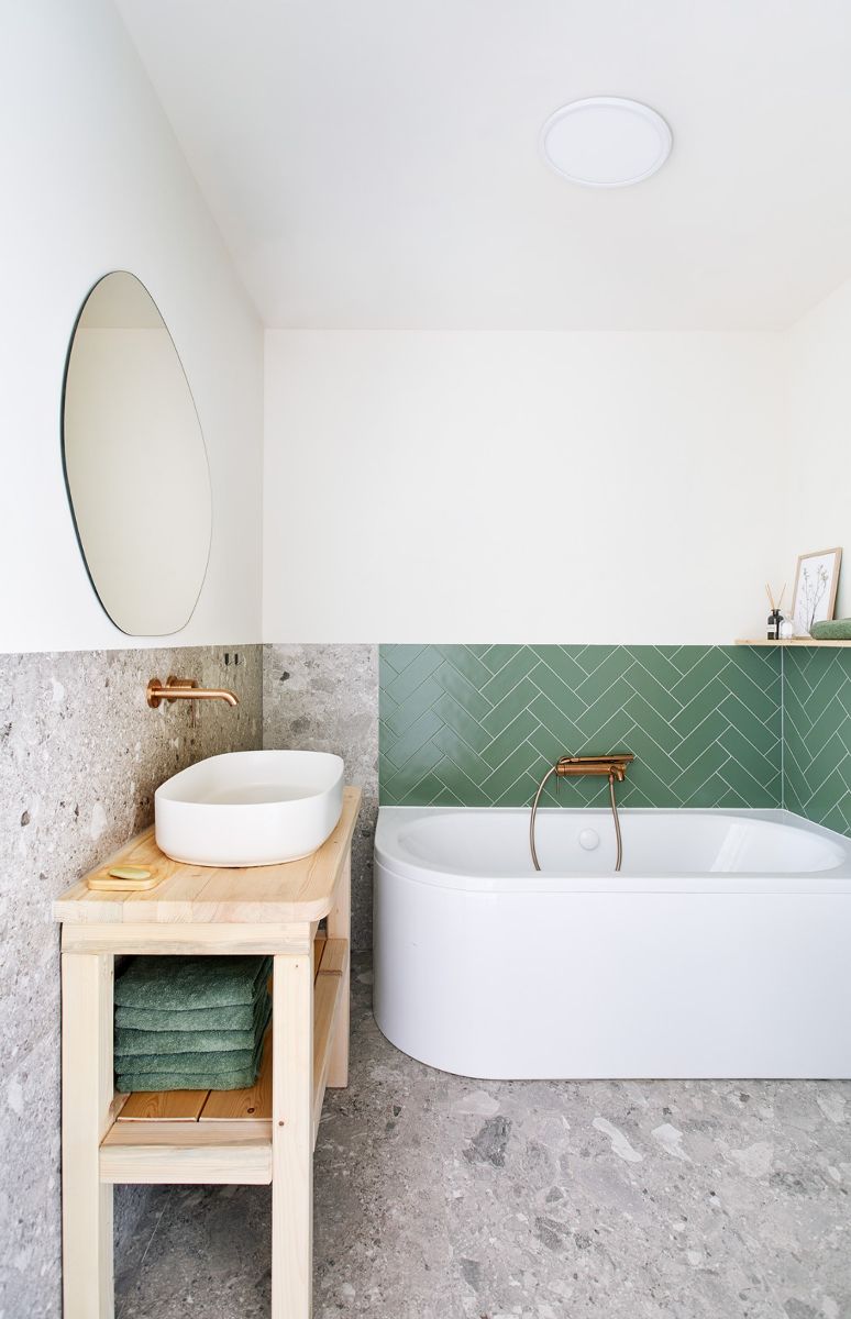 Moderná kúpeľňa s bielou vaňou, zeleným obkladom v tvare rybej kosti a dreveným stolíkom pod oválnym umývadlom.