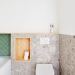 Závesná biela toaleta na stene s terazzovým obkladom a dreveným výklenkom na odkladanie hygienických potrieb.