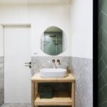 Kúpeľňový kút s bielym keramickým umývadlom, drevenou konzolou a okrúhlym zrkadlom na sivej stene.