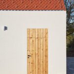 Prírodné drevené dvere, biela štruktúrovaná fasáda, červená pálená škridla a sivý kamenný chodník.