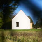 Minimalistický biely dom so strmou sedlovou strechou a úzkym obdĺžnikovým oknom na zelenom trávnatom pozemku.