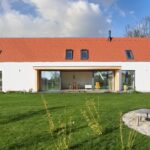 Dlhá biela fasáda s drevenou presklenou terasou, tmavosivými strešnými oknami a vonkajším kamenným ohniskom.