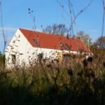 Biely vidiecky dom s tradičnou červenou škridlou a modernými strešnými oknami za divokou lúčnou vegetáciou.